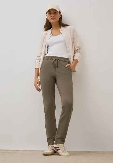 Frau in lässigem Outfit mit Kappe, Cardigan, weißem Top und beigen Jogginghosen.
